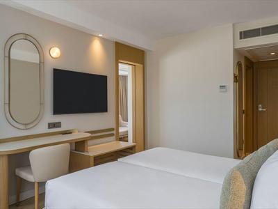 Habitación de hotel luminosa con cama doble, televisor, escritorio y balcón con vista a plantas.