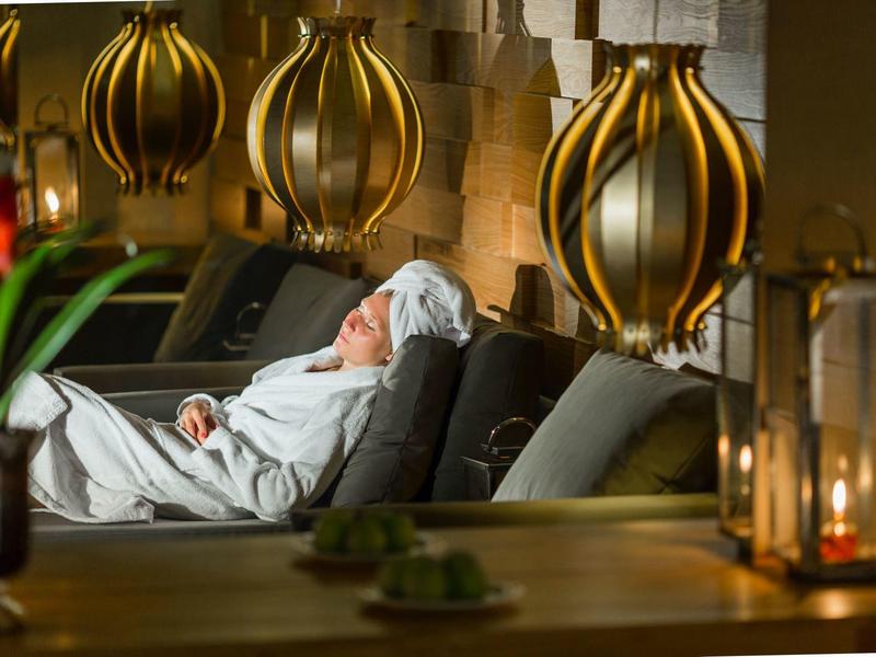 Person relaxes in white bathrobe on lounge chair in cozy indoor setting with golden lamps.