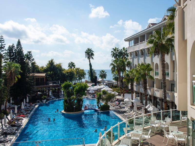 Hotel pool with sun loungers, palm trees, and a view of the sea on a sunny day.