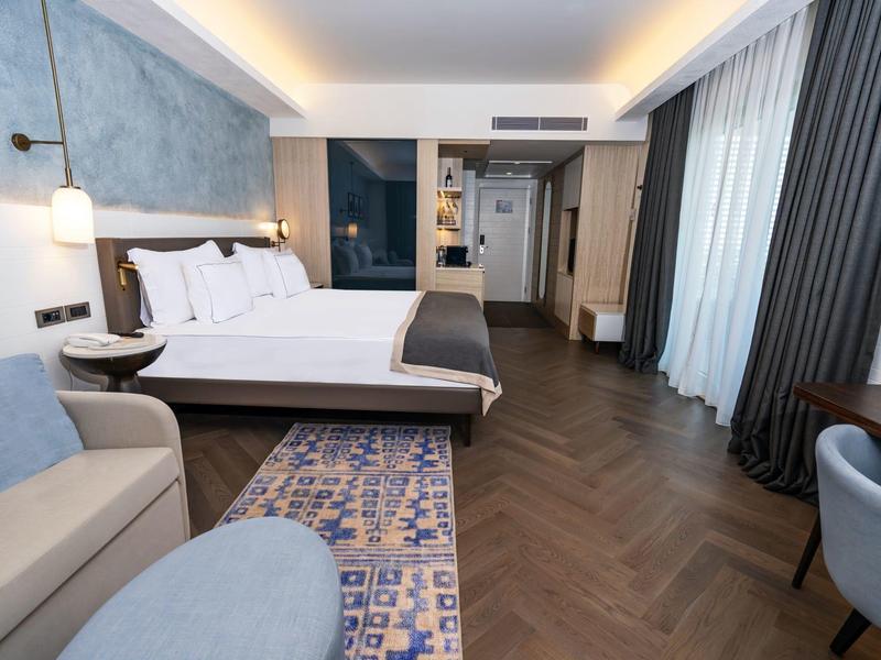 Camera d'hotel moderna con letto matrimoniale, divano, tappeto e tende davanti a finestre a tutta altezza.