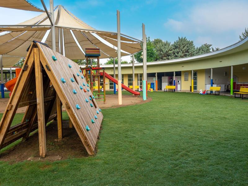 Parco giochi per bambini con parete da arrampicata e altalene colorate sotto tende da sole su prato sintetico.