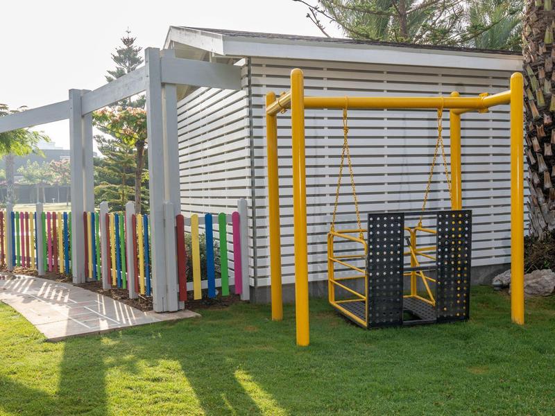 Parco giochi all'aperto con altalena gialla e recinto colorato accanto a un edificio bianco