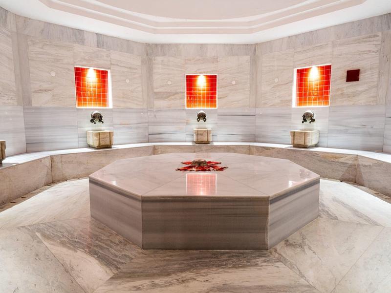 Ambiente hammam moderno e luminoso con fontana centrale in marmo e sedute semicircolari