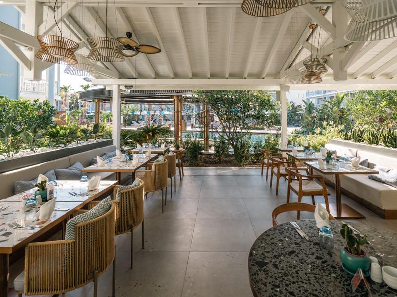 Area esterna luminosa di un ristorante con tavoli in legno, sedie e piante ai lati