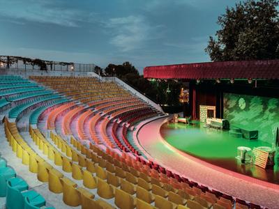 Anfiteatro iluminado con colores, filas de asientos coloridos y escenario al aire libre al anochecer.