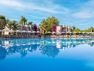Piscina grande con tumbonas, sombrillas y plantas en un resort bajo un cielo azul.