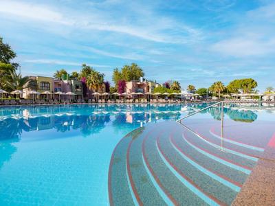 Großer Pool mit Treppenstufen, umgeben von Palmen und Häusern unter blauem Himmel.