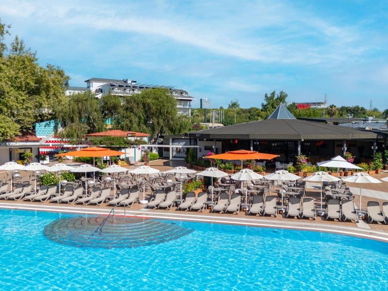 Grande piscina con molti lettini e ombrelloni arancioni davanti a un edificio dell'hotel sotto un cielo blu.