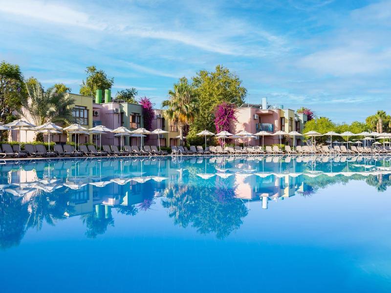 Grande piscina con lettini e palme davanti a edifici moderni di un hotel sotto un cielo azzurro.