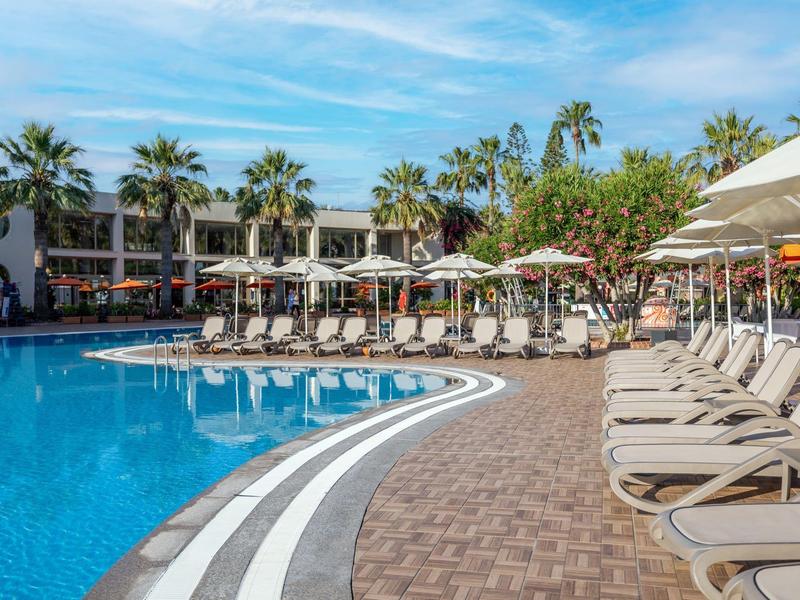 Grande piscina dell'hotel con lettini, ombrelloni e palme in una giornata di sole.