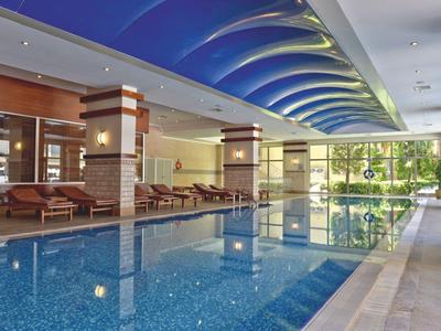 Innenpool mit blauer Deckenbeleuchtung, Liegestühlen und großen Fenstern mit Blick ins Grüne.