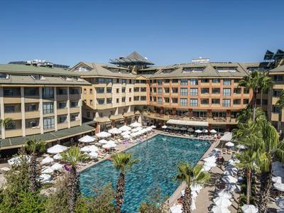 Duży ośrodek z wieloma budynkami, basenem i licznymi leżakami pod niebieskim niebem.