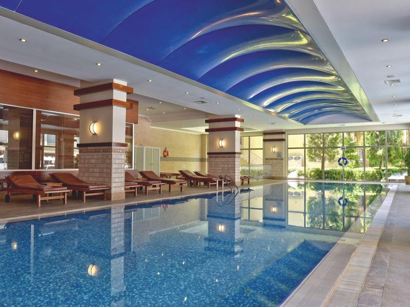 Innenpool mit blauer Deckenbeleuchtung, Liegestühlen und großen Fenstern mit Blick ins Grüne.