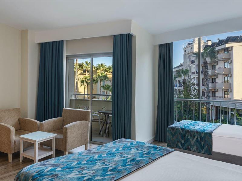 Hotelzimmer mit zwei Betten, Sitzbereich, TV und Blick auf Stadt und Palmen durch große Fenster.