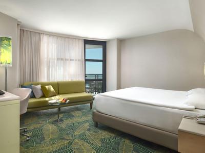 Jasny pokój hotelowy z dużym łóżkiem, zieloną sofą, wykładziną i oknem z widokiem na zewnątrz.