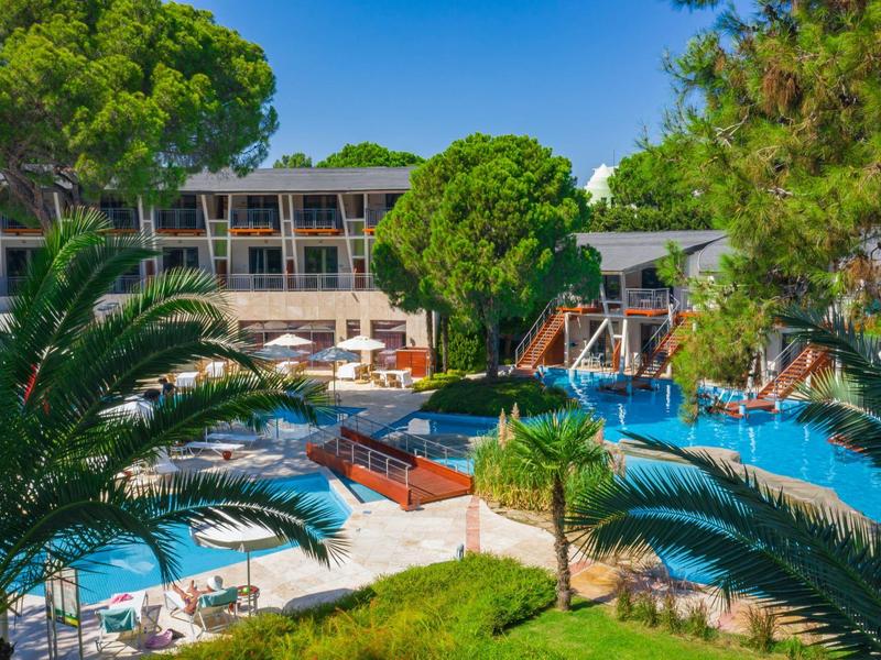 Hotelpool mit Liegestühlen, Palmen und grünem Garten bei klarem Himmel.