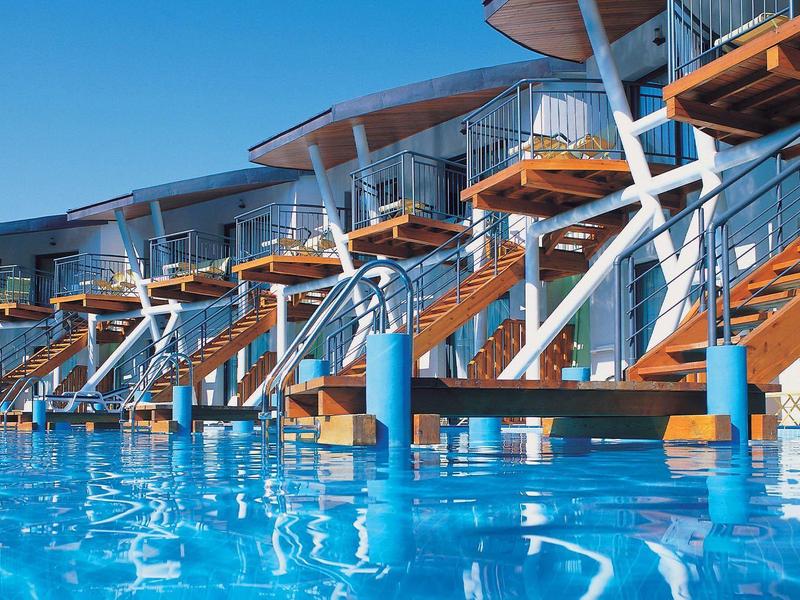 Moderne Hotelanlage mit Holzstegen und Treppen über einem großen Pool bei klarem Himmel.