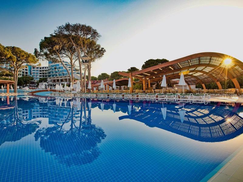 Großer Außenpool mit Sonnenschirmen und modernen Gebäuden im Hintergrund bei Sonnenuntergang.