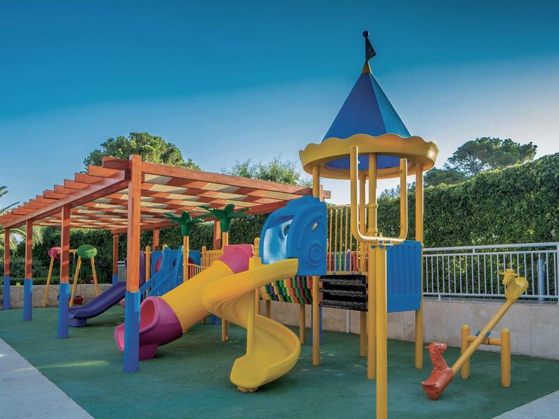 Bunter Spielplatz mit Rutsche und Klettergerüst unter blauem Himmel neben einem schattigen Bereich