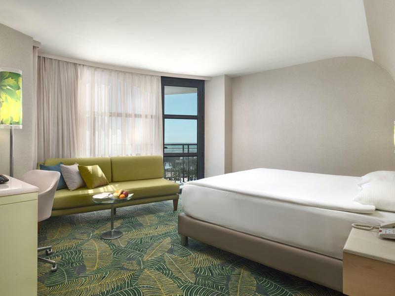 Modernes Hotelzimmer mit Doppelbett, grünem Sofa, Teppichboden und großem Fenster