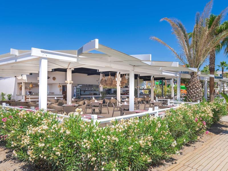 Area esterna di un ristorante con pergolati bianchi, posti a sedere e palme sotto un cielo azzurro brillante