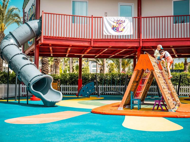 Parco giochi colorato con scivolo e struttura per arrampicata davanti a un hotel con balcone.