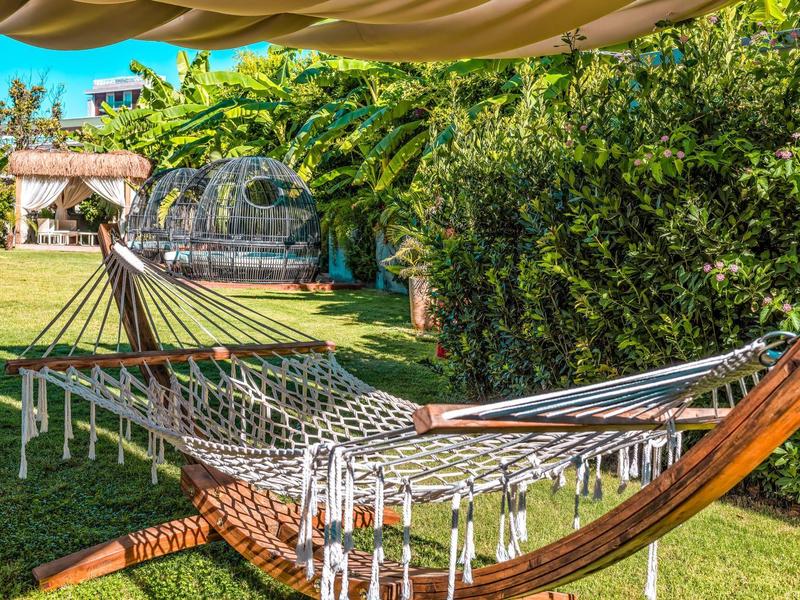 Amaca in giardino con prato verde, cespugli e una serra sullo sfondo.