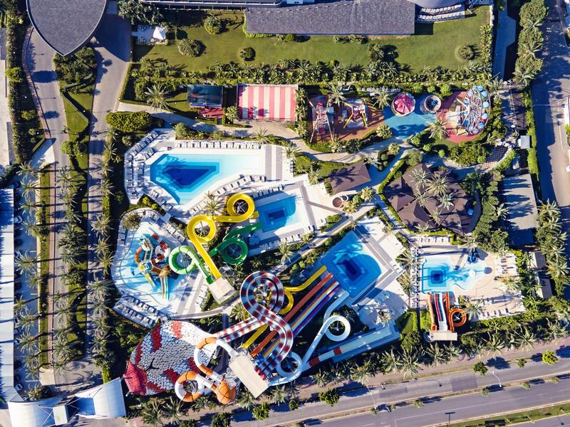 Vista aérea de un parque acuático con varias piscinas, toboganes y áreas verdes junto a una carretera.