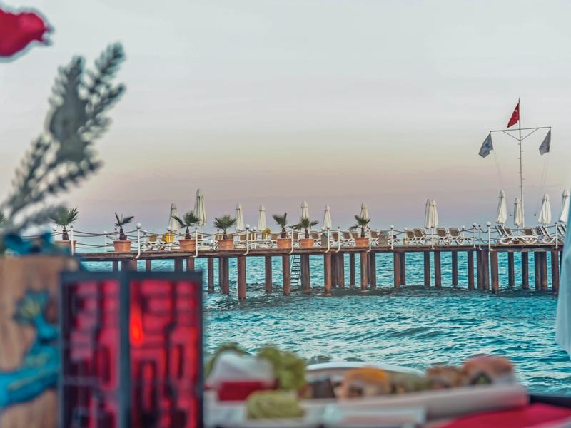 Vista de un muelle con mesas y sillas junto al mar con tiempo tranquilo y veleros al fondo.