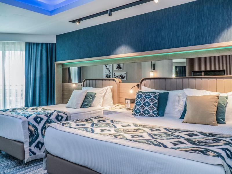 Habitación moderna de hotel con dos camas grandes, acentos azules y luz natural exterior.