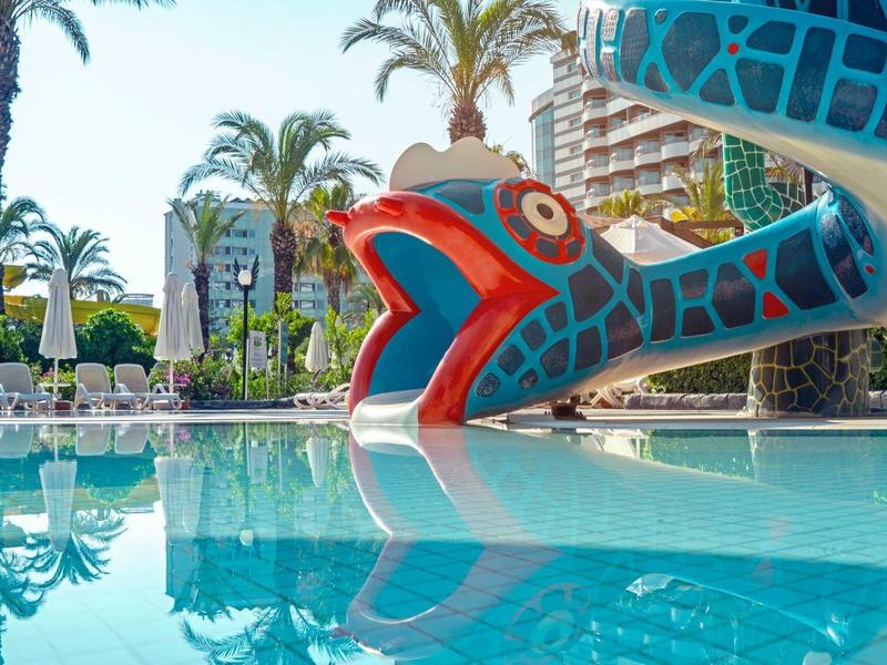 Boca colorida de tobogán acuático en piscina de hotel con palmeras y tumbonas de fondo.