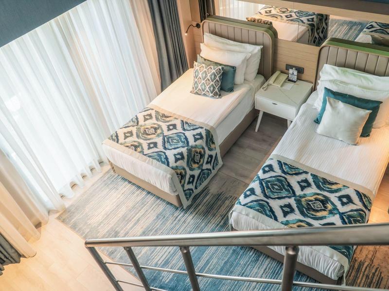 Dos camas individuales en una habitación de hotel luminosa con mantas y alfombra estampadas.