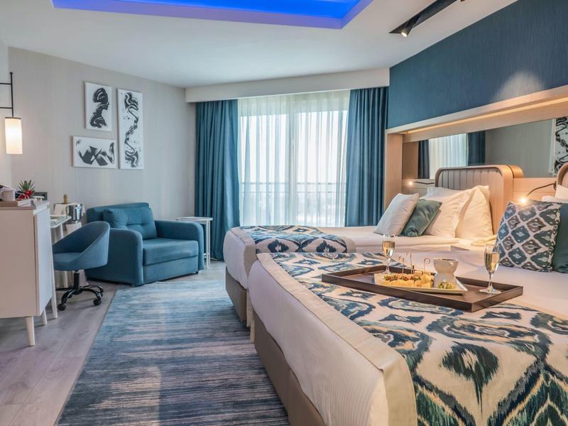 Habitación de hotel moderna con cama grande, sillones azules y ventana con cortinas.