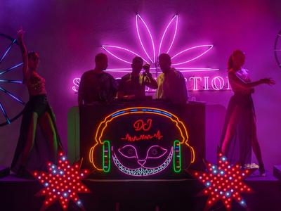 DJ dietro una consolle illuminata con orecchie da coniglio al neon e ballerini in luce viola.