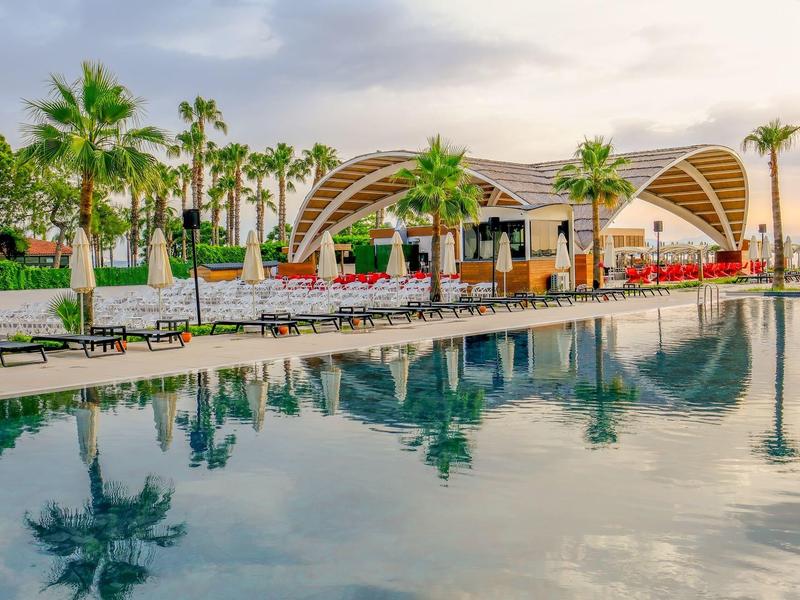 Modern pavilion by hotel pool with palm trees and calm water at sunset.