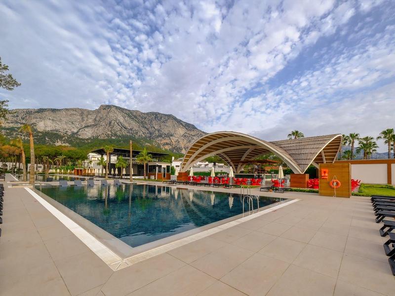 Modern pool with sun loungers, pavilion, and mountains in the background under cloudy sky.