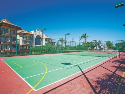 Prázdný tenisový kurt vedle hotelu s modrou oblohou a palmami v pozadí.