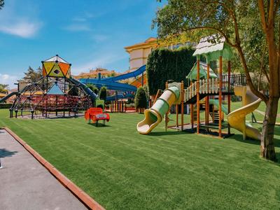 Speelplaats voor kinderen met glijbanen en klimtoestellen op groen kunstgras bij zonnig weer.