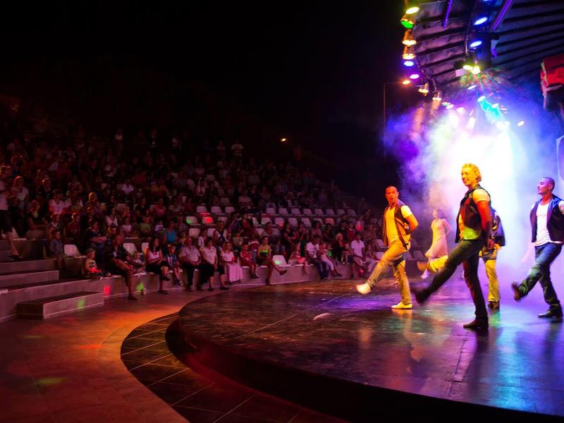 Palco illuminato con tre ballerini di fronte a un vasto pubblico in un teatro.