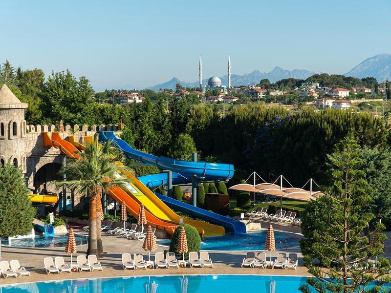 Area con scivoli d'acqua colorati accanto alla piscina, palme e montagne sullo sfondo.