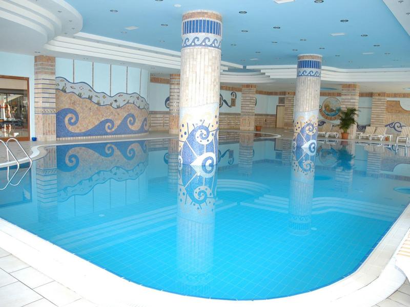 Piscina interna con acqua blu, pavimento piastrellato bianco e colonne decorate.