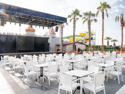 Área al aire libre con mesas y sillas blancas, escenario y palmeras junto a la piscina del hotel.