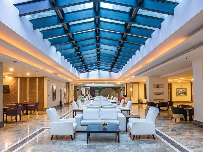 Área de lounge luminosa del hotel con sillones blancos, techo de vidrio azul y iluminación elegante.