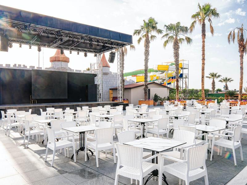 Área al aire libre con mesas y sillas blancas, escenario y palmeras junto a la piscina del hotel.
