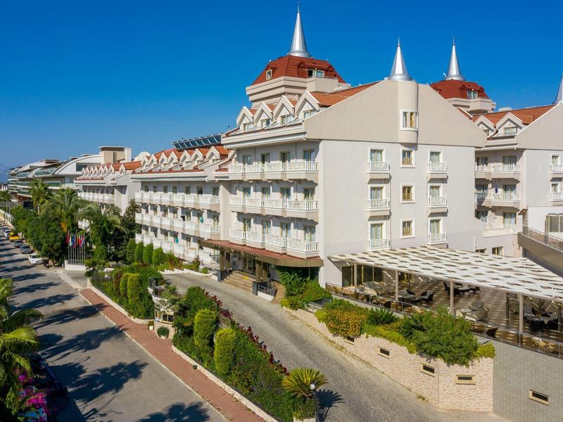 Gran edificio de hotel con balcones blancos junto a una calle arbolada bajo un cielo azul claro.