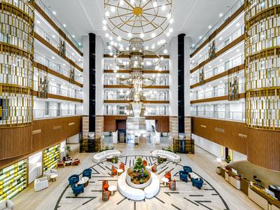 Jasna, nowoczesna recepcja hotelowa z wysokimi ścianami, złotymi lampami i miejscami do siedzenia na marmurowej podłodze.