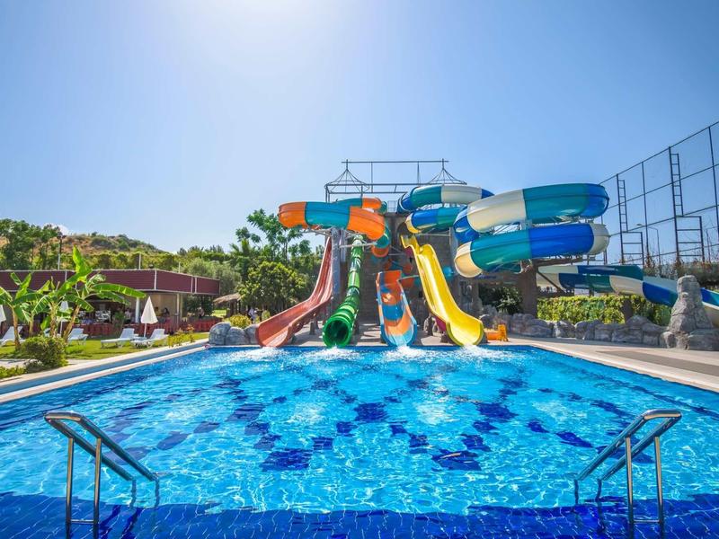 Barevný vodní park s průzračným bazénem a slunečnou oblohou, obklopený palmami.