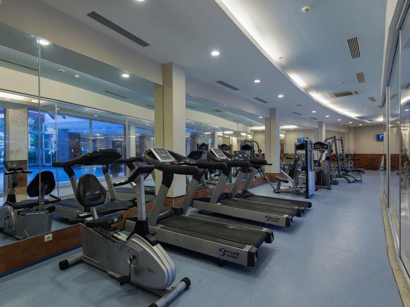 Hotelová posilovna s běžeckými pásy a jízdními koly, zrcadlová stěna a velké okno.