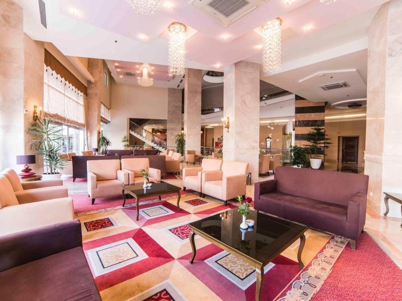 Světlý hotelový salonek s barevným kobercem, několika sezeními a velkými okny.