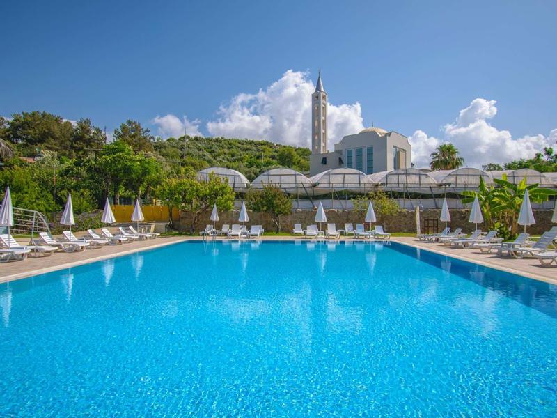 Velký hotelový bazén s bílými lehátky a slunečníky před zelenými kopci a modrou oblohou.